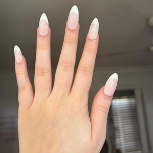 May include: A hand displaying long, oval nails. Each nail has a pale pink base and a white tip, in a classic French manicure design. The nails are smooth and glossy, reflecting light.