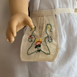 May include: A white doll dress with a cream-colored embroidered apron. The apron features a floral design with green stems and colorful flower details. A cream-colored woven belt cinches the waist.