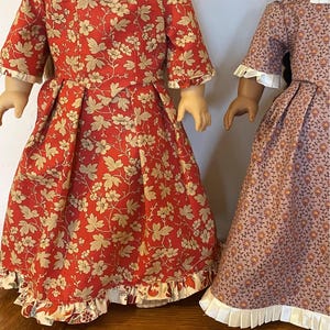 Puede incluir: Dos muñecas con vestidos de estampado floral. La muñeca de la izquierda lleva un vestido rojo con un ribete de encaje blanco. La muñeca de la derecha lleva un vestido rosa con un ribete de encaje blanco.