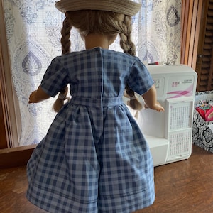 Peut inclure: Une poupée portant une robe en vichy bleu et blanc avec un chapeau de paille. La poupée a les cheveux blonds en tresses et se tient devant une machine à coudre.