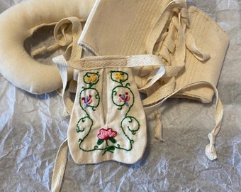 Könnte beinhalten: Ein Set aus weißen Stoffpuppenkleidern mit gestickten Blumendetails. Das Set beinhaltet ein Mieder, einen Rock und ein Paar weiße Stoffbänder.