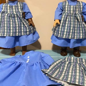 May include: Two dolls wearing blue dresses with a plaid apron and white buttons. The dolls are standing on a light blue surface. There is a third dress on the surface, also blue with a plaid apron.