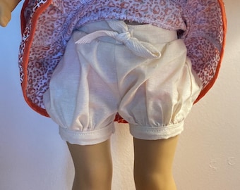 Puede incluir: Una muñeca con un vestido de estampado floral blanco y rojo con un volante blanco y pantalones cortos blancos y abullonados. La muñeca lleva chanclas de rayas blancas y rosas.