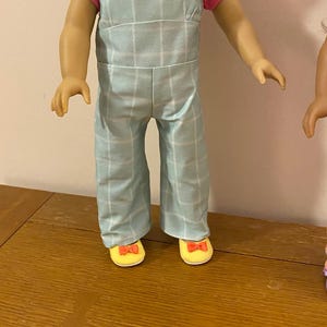 May include: A doll wearing a light blue and white plaid overall dress with pink buttons and a pink short-sleeved shirt. The doll is wearing yellow shoes with a red bow.