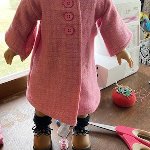 May include: A pink knitted doll coat with four pink buttons and a blue and white checked collar.