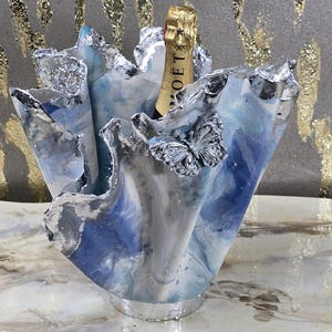 May include: A blue and white resin champagne bottle holder with a silver swirl design. The holder is shaped like a flower and has a gold bottle inside.