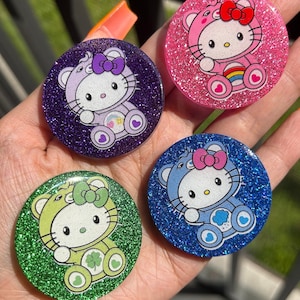 May include: Four round phone grips with glitter backgrounds and Hello Kitty wearing different colored outfits. The outfits are a rainbow, a cloud, a shamrock, and a purple heart.