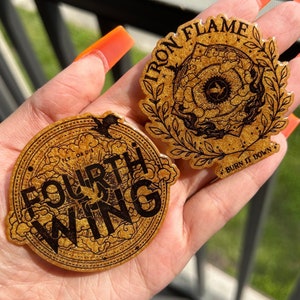 May include: Two gold glitter stickers with black text. The first sticker says "Fourth Wing" with a bird design. The second sticker says "Iron Flame" with a stylized eye design and the text "Burn it down".