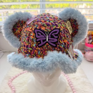 May include: A crochet bucket hat featuring a multi-coloured yarn base of purple, orange, and green. The hat has light blue faux fur trim along the brim and two large faux fur ears. A prominent purple and black butterfly patch is centred on the front.
