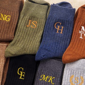 May include: A collection of ribbed socks in various colours including brown, olive green, navy blue, black, grey, and tan. Each sock features embroidered initials or names such as NG, JS, GH, MICHAEL, M|G, GE, MK, and S in yellow or white thread.