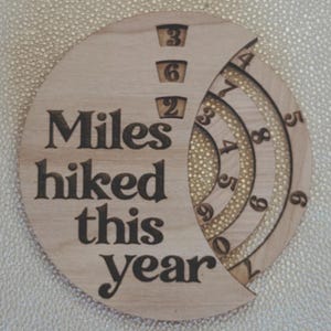 Wooden circular plaque with the words "Miles hiked this year" engraved in black. The plaque has a section with numbers and a series of concentric rings, and a section with the numbers 1, 2, and 9