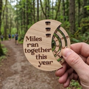 May include: A circular light-colored wooden tracker with the engraved text "Miles ran together this year" and three rotating rings featuring numbers zero through nine, allowing for a manual count of miles run throughout the calendar year.
