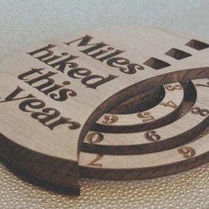 Wooden circular plaque with the words "Miles hiked this year" engraved in black. The plaque has a section with numbers and a series of concentric rings, and a section with the numbers 1, 2, and 9