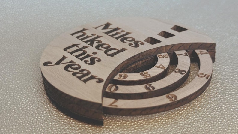 Wooden circular plaque with the words "Miles hiked this year" engraved in black. The plaque has a section with numbers and a series of concentric rings, and a section with the numbers 1, 2, and 9