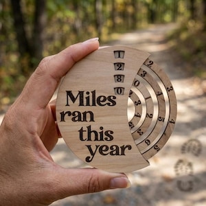 May include: A wooden running tracker with the text "Miles ran this year" is held up in front of a blurred outdoor background. The tracker has a circular design with concentric rings and numbers to track mileage.
