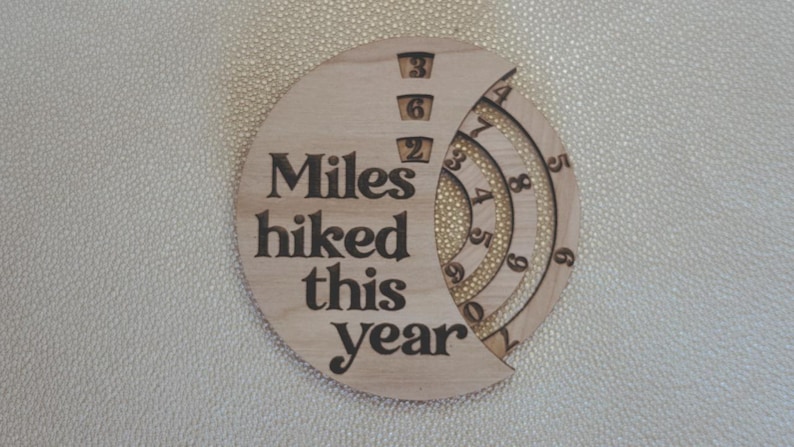 Wooden circular plaque with the words "Miles hiked this year" engraved in black. The plaque has a section with numbers and a series of concentric rings, and a section with the numbers 1, 2, and 9