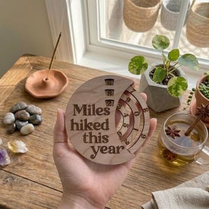 May include: A wooden disc with the text "Miles hiked this year" engraved on it. The disc has a rotating dial to track mileage. The image also shows a small potted plant, crystals, and a cup of tea.