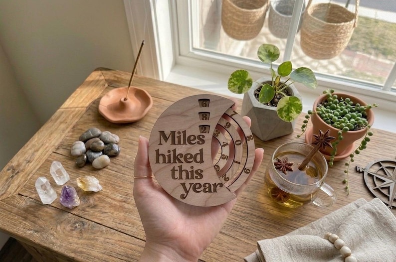 May include: A wooden disc with the text "Miles hiked this year" engraved on it. The disc has a rotating dial to track mileage. The image also shows a small potted plant, crystals, and a cup of tea.