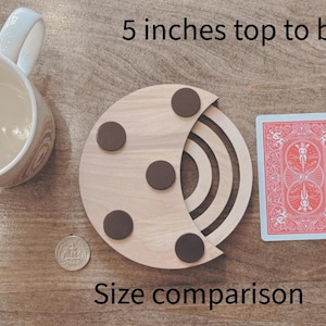 May include: A wooden coaster with a crescent moon design, featuring four brown circular accents. A playing card, a pencil, a mug, and a coin are next to the coaster for size comparison. The text "5 inches top to bottom" and "Size comparison" are visible.