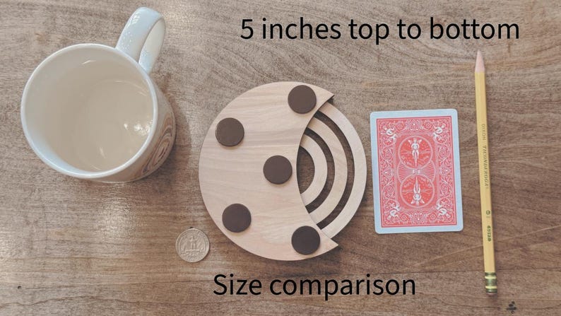 May include: A wooden coaster with a crescent moon design, featuring four brown circular accents. A playing card, a pencil, a mug, and a coin are next to the coaster for size comparison. The text "5 inches top to bottom" and "Size comparison" are visible.