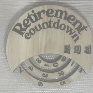 May include: A circular light-colored wooden retirement countdown plaque featuring the text "Retirement countdown" at the top. The plaque includes three rotating rings with printed numbers and a fixed display window showing the digits 8, 0, and 3.