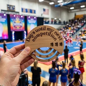 May include: A circular wooden countdown plaque featuring the engraved text "Days until my next competition" with three rotating inner rings displaying numbers to track the remaining days, held against a blurred indoor sports arena background.