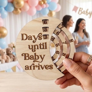 May include: A circular wooden countdown sign with the engraved text Days until Baby arrives. The sign features a crescent-shaped cutout with rotating inner rings displaying numbers to track the remaining time until a baby is born.