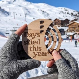 Peut inclure: Un compteur circulaire en bois avec le texte gravé "Days skied this year" comprenant trois anneaux intérieurs rotatifs avec des chiffres pour compter et afficher manuellement le nombre total de jours passés à skier durant la saison.