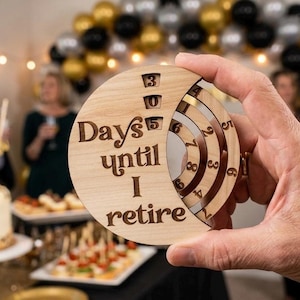 May include: A wooden retirement countdown disc with the text "Days until I retire" and a dial showing "305" days. The disc is held in a hand, with a blurred party scene in the background, including a cake and balloons.