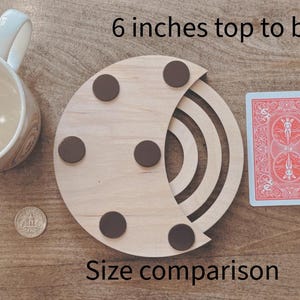 May include: A wooden coaster with a crescent design, featuring brown circular pads. The image includes a playing card, a pencil, a coffee mug, and a coin for size comparison. The coaster is 6 inches from top to bottom. The text "Size comparison" is also visible.
