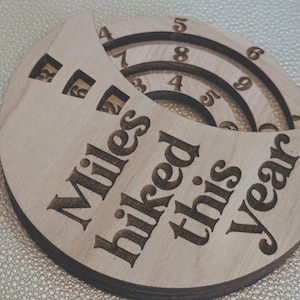 Wooden circular plaque with the words "Miles hiked this year" engraved in black. The plaque has a section with numbers and a series of concentric rings, and a section with the numbers 1, 2, and 9