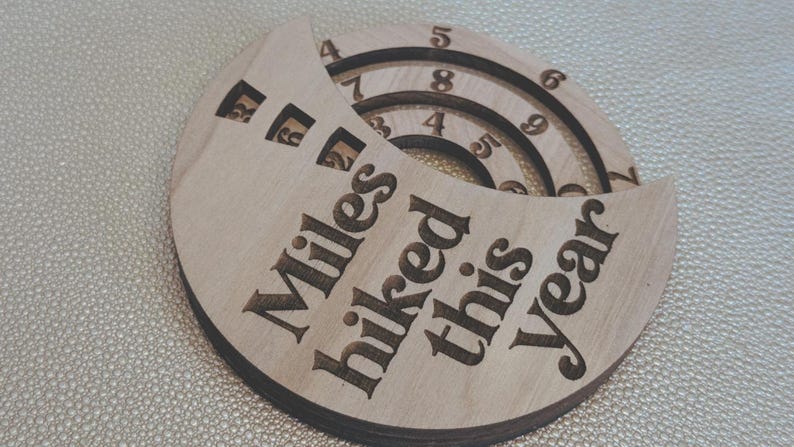 Wooden circular plaque with the words "Miles hiked this year" engraved in black. The plaque has a section with numbers and a series of concentric rings, and a section with the numbers 1, 2, and 9