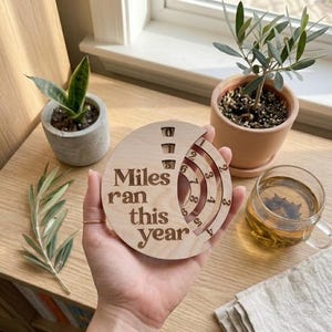 May include: A wooden disc with the text "Miles ran this year" is held in a hand. The disc has a circular design with carved markings for tracking miles. The background includes plants, a cup of tea, and a window.