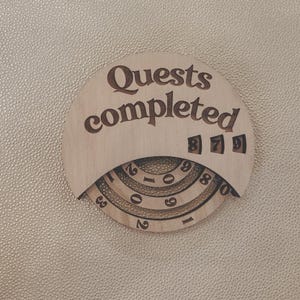 May include: A circular light-colored wooden tracker with the text "Quests completed" engraved at the top. It features three rotating concentric rings with numbers zero through nine, and three small windows displaying the digits eight, seven, and nine.