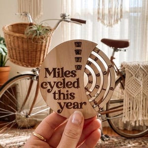May include: A circular light-colored wooden tracker with the engraved text Miles cycled this year. The tracker features four concentric rotating rings with numbers zero through nine, allowing for manual adjustment to record cycling progress.