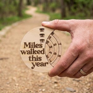 May include: A circular light-coloured wooden tracker with the engraved text Miles walked this year. The device features rotating concentric rings with numbers to manually record distance, including a vertical column showing the digits 1, 3, and 5.