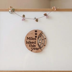 May include: Wooden wall decor with the text "Miles hiked this year" engraved on a circular design. The design has a rotating dial to track hiking progress. The decor is mounted on a white background within a wooden frame.