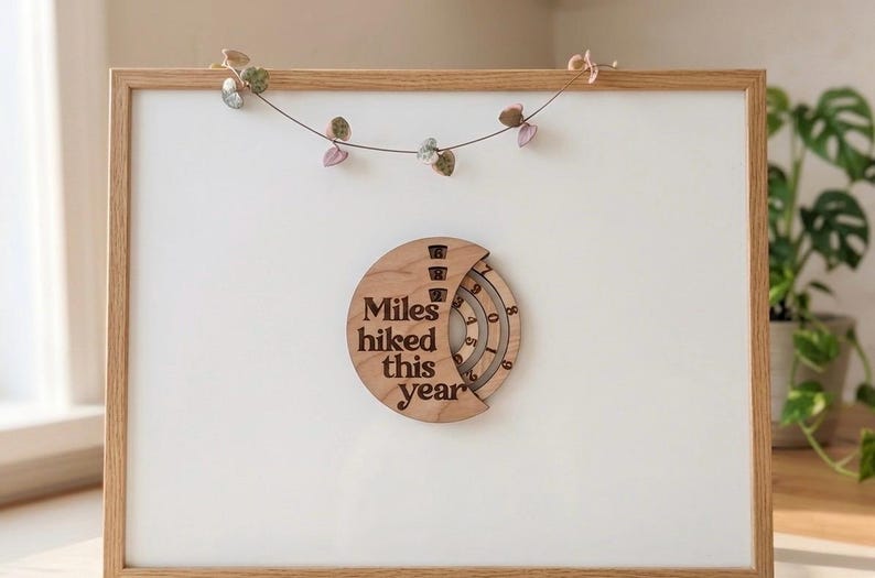 May include: Wooden wall decor with the text "Miles hiked this year" engraved on a circular design. The design has a rotating dial to track hiking progress. The decor is mounted on a white background within a wooden frame.
