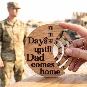 May include: A circular wooden countdown calendar featuring the engraved text "Days until Dad comes home" with three rotating inner rings displaying numbers to track time, constructed from light brown wood with dark brown lettering.