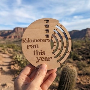 May include: A circular light-colored wooden tracker with the text "Kilometers ran this year" engraved on the left side. The right side features concentric rings with numbers zero through nine, allowing for manual adjustment to track running distance.