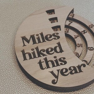 Wooden circular plaque with the words "Miles hiked this year" engraved in black. The plaque has a section with numbers and a series of concentric rings, and a section with the numbers 1, 2, and 9