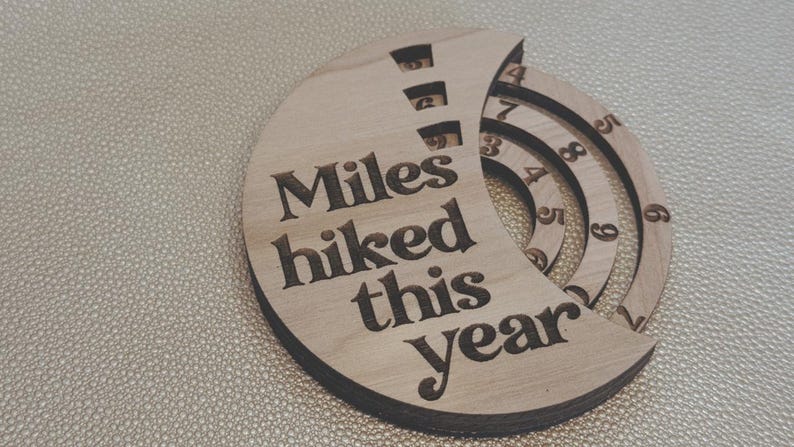 Wooden circular plaque with the words "Miles hiked this year" engraved in black. The plaque has a section with numbers and a series of concentric rings, and a section with the numbers 1, 2, and 9