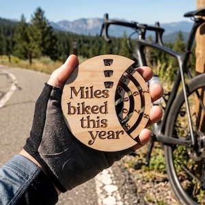 May include: A wooden cycling tracker with the text "Miles biked this year" is held up in front of a bicycle. The tracker has a circular design with a rotating dial to track mileage. The background shows a paved path and trees.