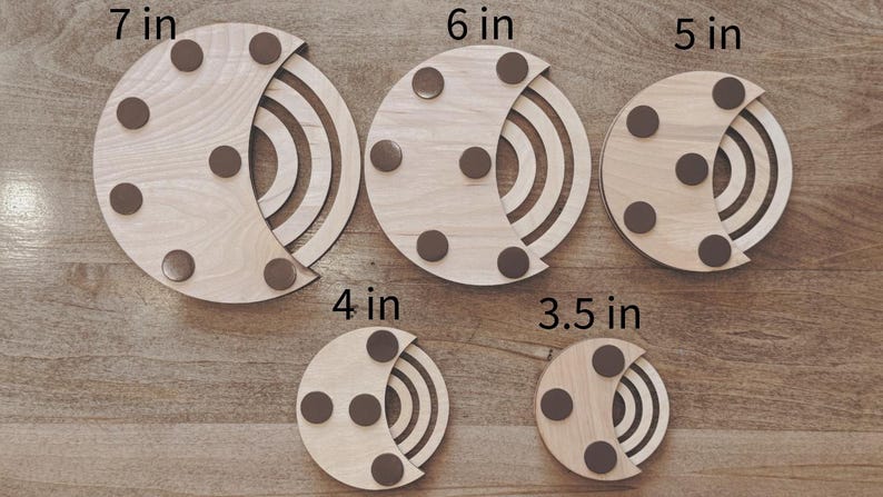May include: A collection of five wooden coasters in varying sizes, ranging from 3.5 inches to 7 inches in diameter. Each coaster features a light wood finish, a crescent-shaped design, and brown circular pads.