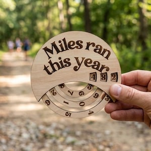 May include: A circular light-colored wooden tracker with the engraved text "Miles ran this year" and three rotating numbered wheels displaying the digits 4, 8, and 2, designed to manually record running progress with a natural wood grain finish.