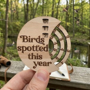 May include: A wooden bird-watching tracker with the text "Birds spotted this year" engraved on it. The tracker has a rotating dial to record the number of birds seen. Binoculars and a field guide are also visible.