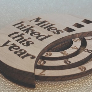 Wooden circular plaque with the words "Miles hiked this year" engraved in black. The plaque has a section with numbers and a series of concentric rings, and a section with the numbers 1, 2, and 9
