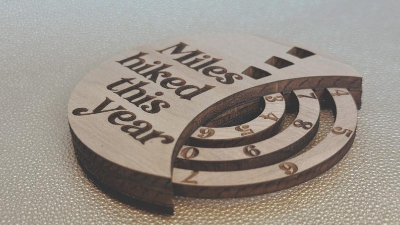 Wooden circular plaque with the words "Miles hiked this year" engraved in black. The plaque has a section with numbers and a series of concentric rings, and a section with the numbers 1, 2, and 9