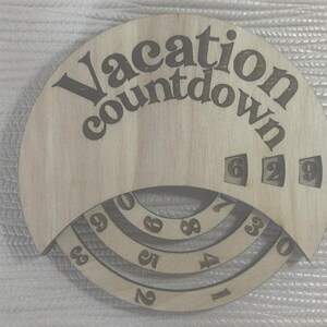 May include: A circular light-colored wooden vacation countdown sign featuring the text "Vacation countdown" engraved at the top. The sign includes three rotating concentric rings with numbers zero through nine to track days, showing the digits 6, 2, and 9.