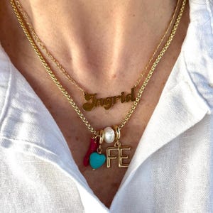 May include: A layered gold necklace with the name "Ingrid" in cursive script. The lower chain features charms, including a turquoise heart, a pearl, and the letters "FE". The necklace is worn over a white shirt.